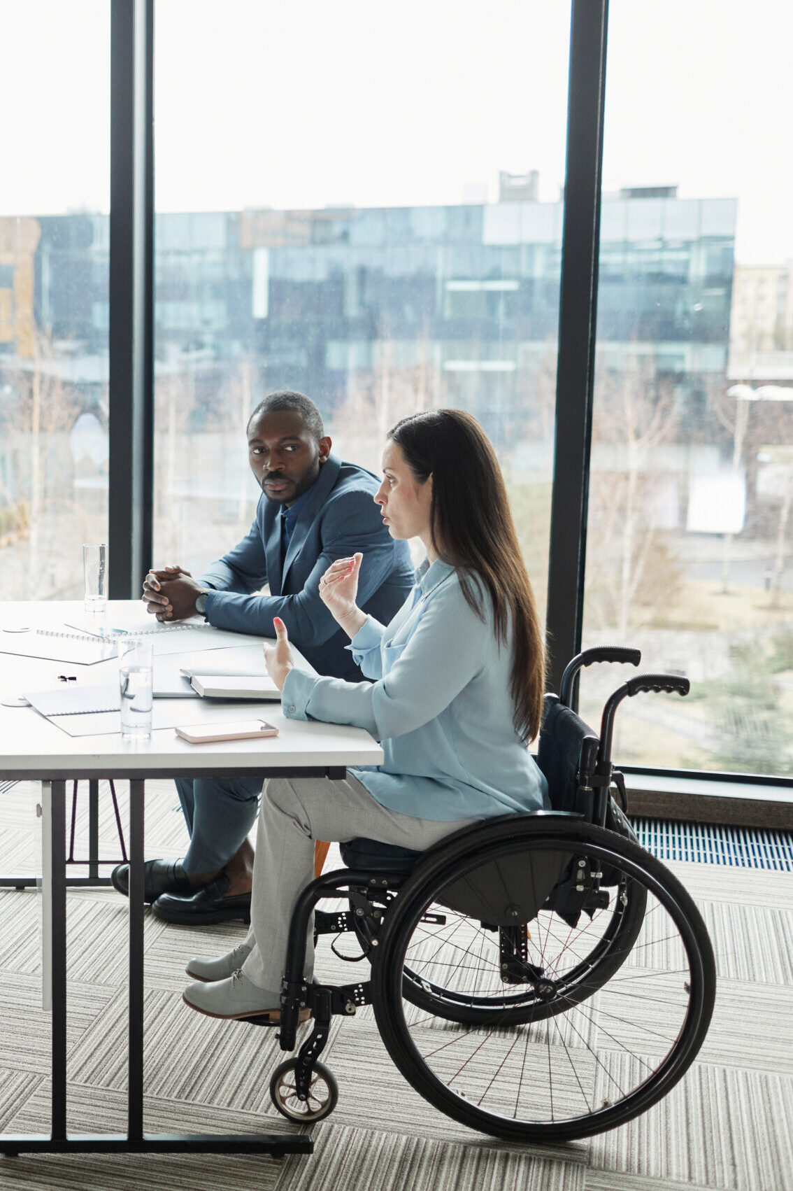 Primrose Watson Implementing diversity Diverse business team discussing project at table in modern office with focus on young woman in wheelchair sharing ideas in foreground.